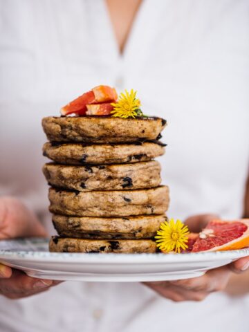 一名女子拿着一堆无蛋煎饼，盘子里有巧克力片，上面有鲜花和葡萄柚角。
