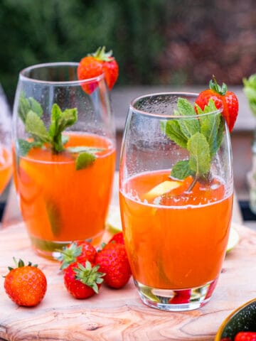 Strawberry lemonade in two tall glasses garnished with mint leaves and strawberries on a wooden board with an outside background.