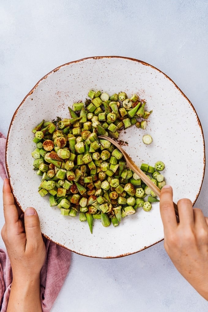 Oven roasted Okra Recipe is for those who hate okra. These crispy roasted okra chips will become their new favorite snack. Healthy and incredibly tasty! #roastedokra #okrarecipe #healthysnack 用木勺将秋葵碎和香料放入白色陶瓷碗中搅拌。
