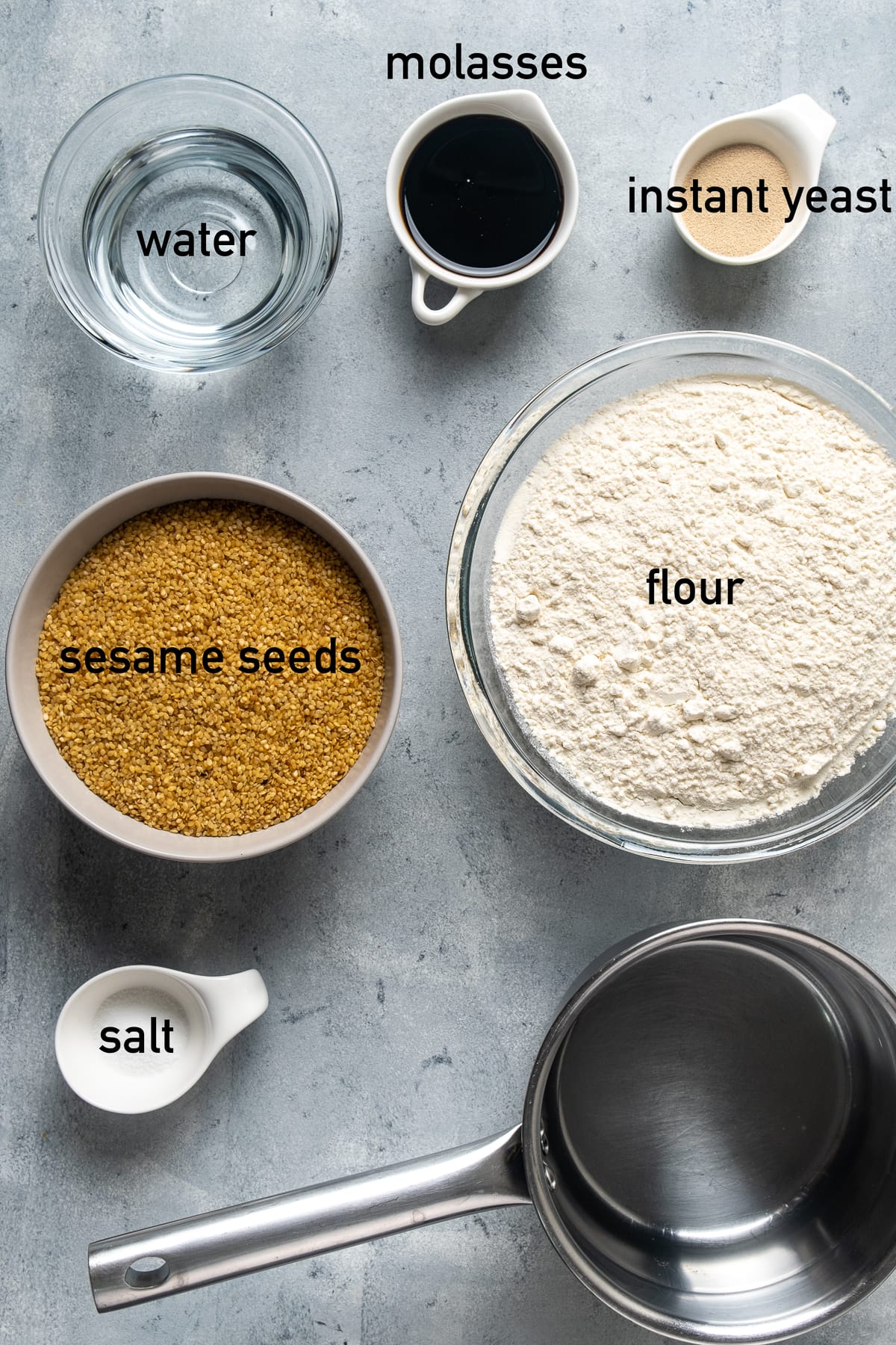 芝麻籽、面粉、葡萄糖蜜、速溶酵母、水和盐都放在不同的碗里，放在灰色的背景上。