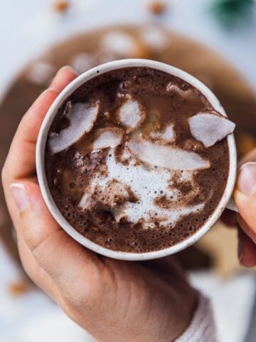 Holding a cup of almond milk hot chocolate topped with coconut oil and coconut flakes.