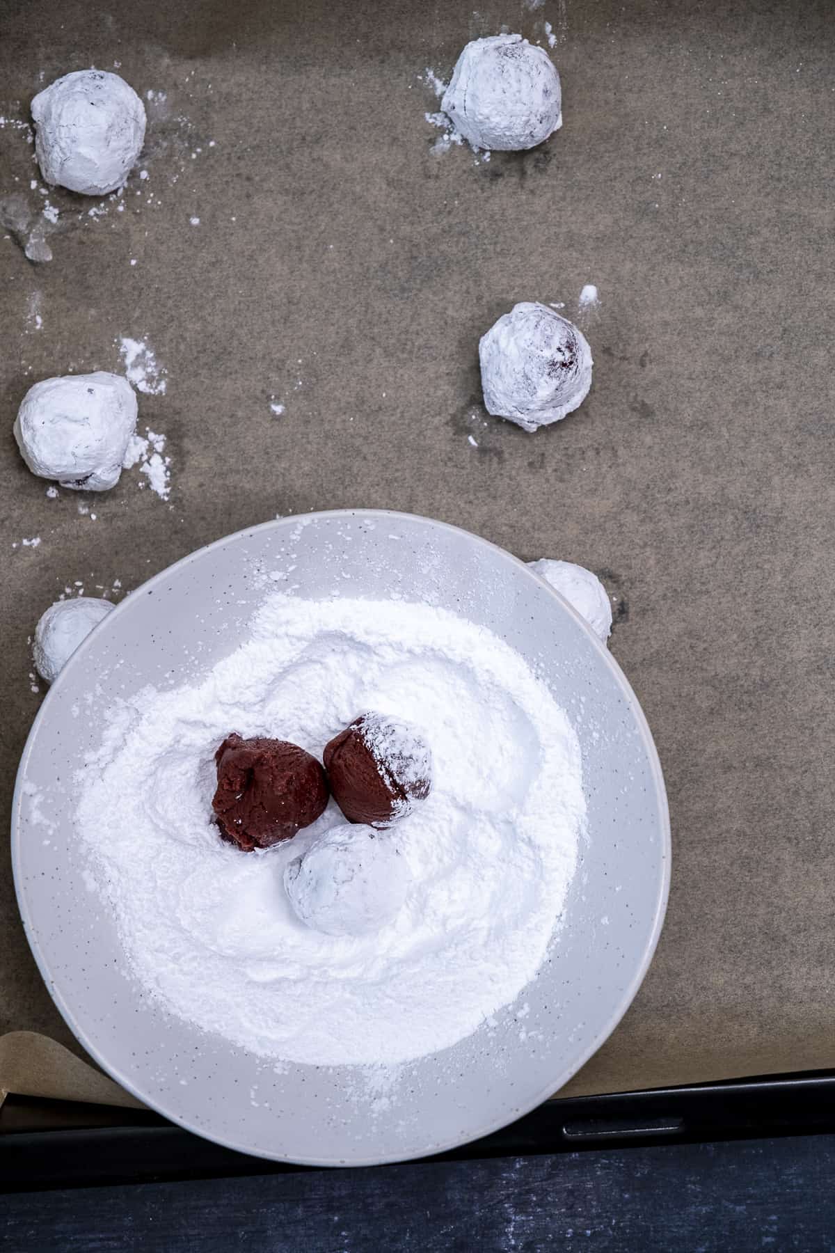 在一碗糖粉里放上红丝绒曲奇面团球，在烤盘上放上更多的曲奇面团球。