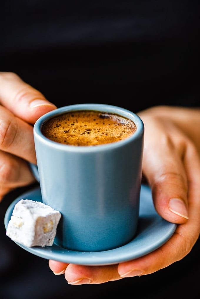 穿着黑色毛衣的女士拿着灰色咖啡杯里的土耳其咖啡，旁边放着开心果土耳其软糖。