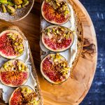 Two bread slices topped with cream cheese and fresh figs dipped coated with tahini and crumbled pistachios on a round wooden board.