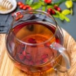 在一个玻璃茶壶的新鲜的玫瑰果茶在一个木板，新鲜的野玫瑰果和蜂蜜浸染料在边。