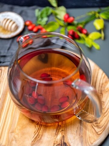 新鲜的玫瑰果茶装在玻璃茶壶里放在木板上，旁边放着新鲜的玫瑰果和蜂蜜。