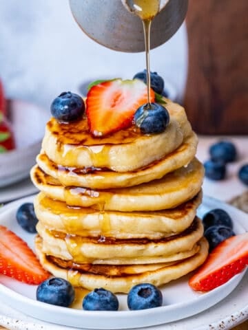 Maple syrup being poured over a stack of pancakes garnished with blueberries and strawberries.