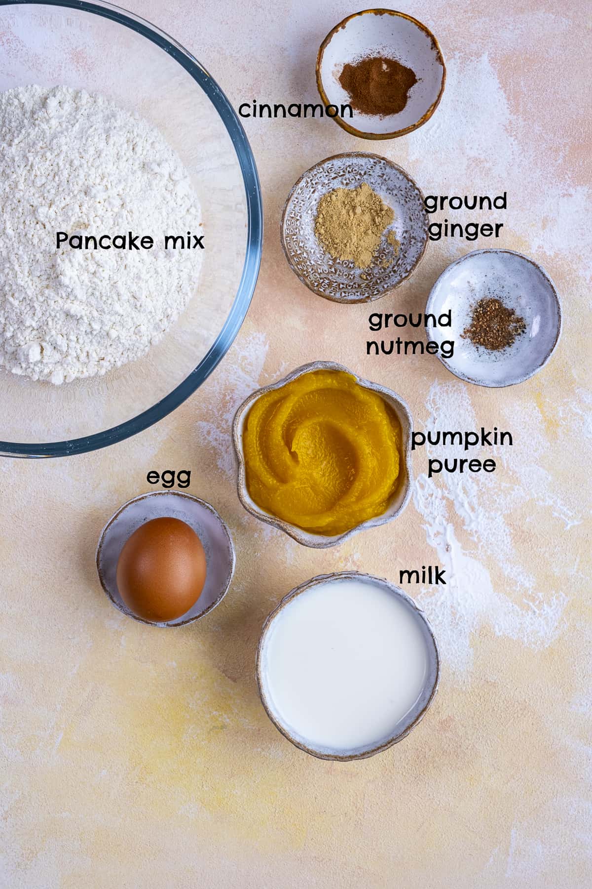 把煎饼粉、肉桂、姜、肉豆蔻、南瓜泥、鸡蛋和牛奶分别放在不同的碗里，放在浅色的背景上。