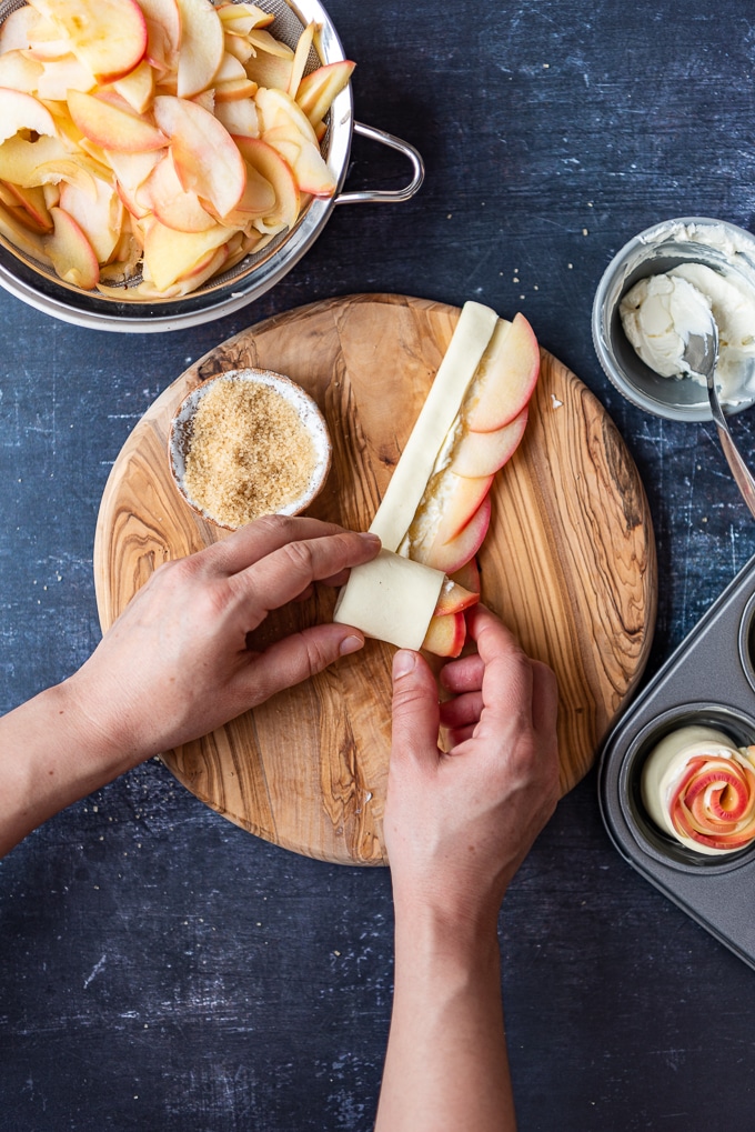 在一个圆形的木板上，用手擀出一个充满苹果片和奶油奶酪的酥皮条。