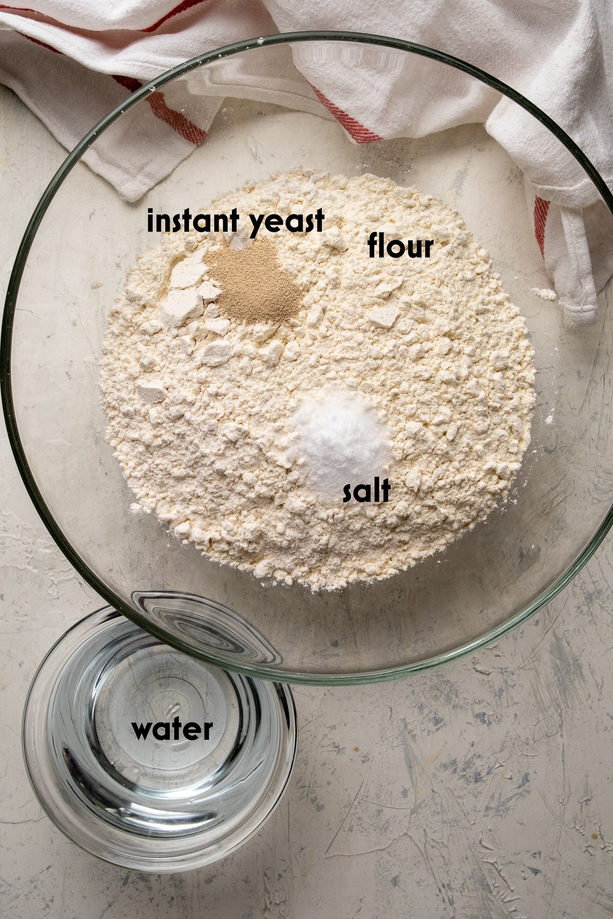 面粉，即食酵母和盐在一个大的玻璃碗里，水在一个小碗里，在浅色的背景上。