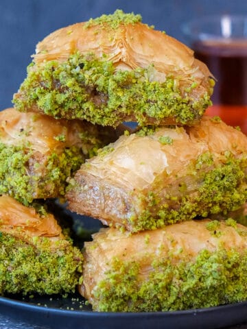 A close shot of Turkish baklava with ground pistachios piled on a plate and a glass of Turkish tea behind these.