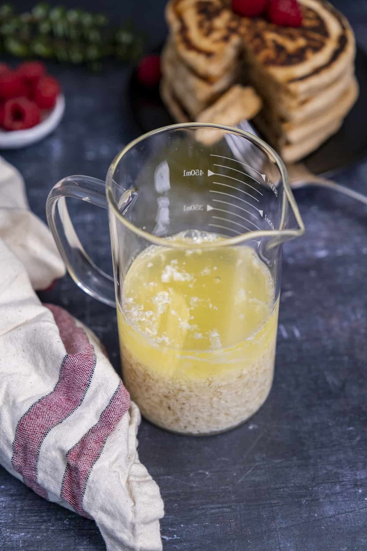 杏仁酪乳装在玻璃杯里，米色亚麻布边有红色条纹，后面是覆盆子和煎饼。