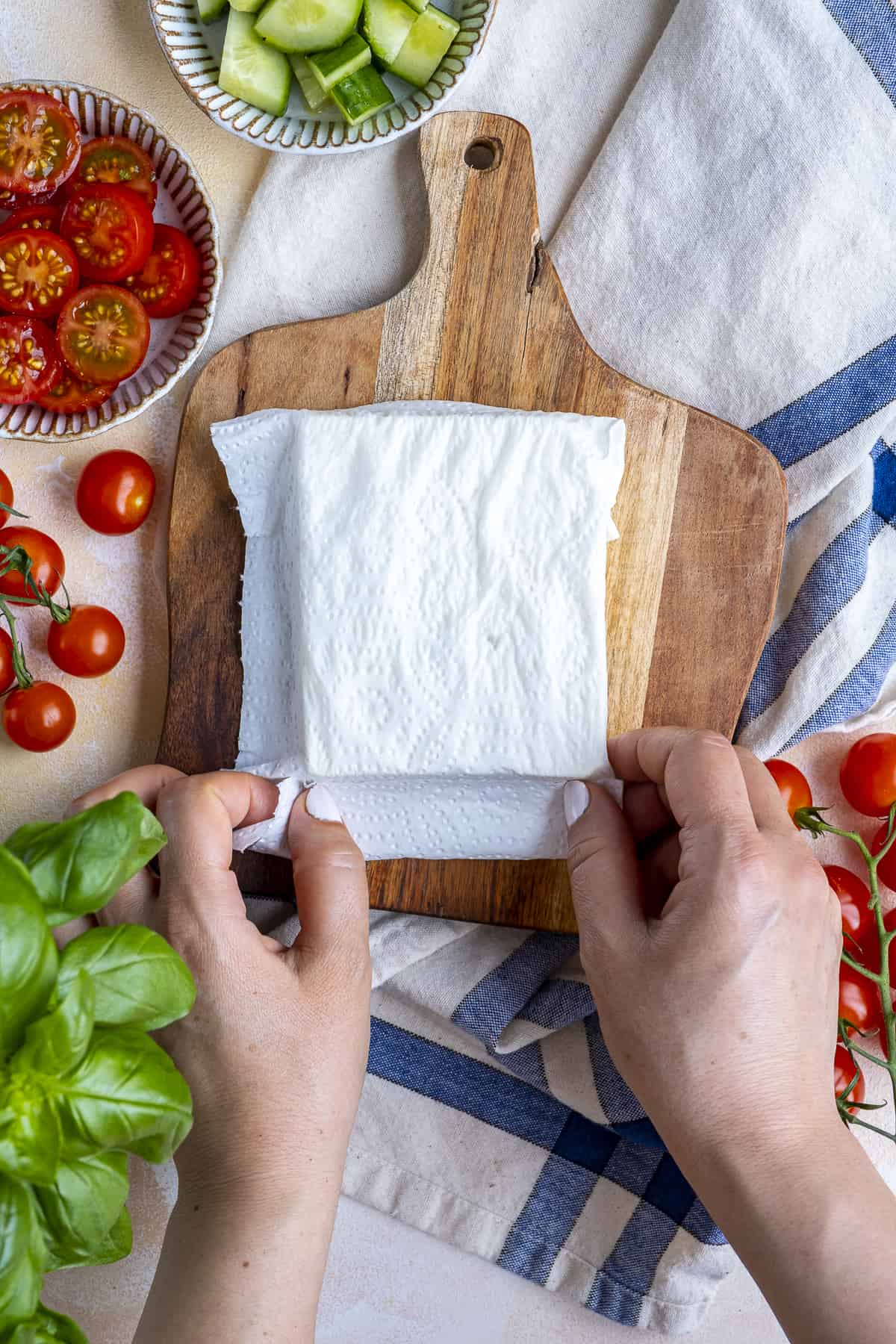 双手用湿纸巾包着一块菲达奶酪。切好的西红柿，黄瓜和新鲜的罗勒。